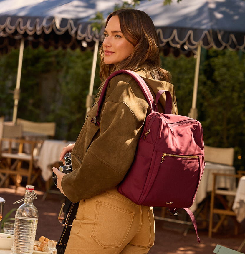 Woman outdoors wearing a burgundy Baggallini anti-theft backpack with gold hardware.