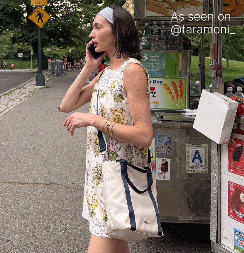 Woman walking on a city sidewalk while talking on her phone and carrying a cream tote bag with dark trim.