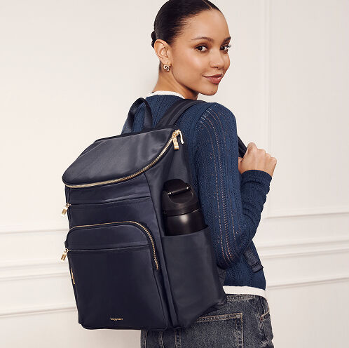 Model wearing a navy Baggallini nylon backpack with gold-tone hardware and side water bottle pocket.