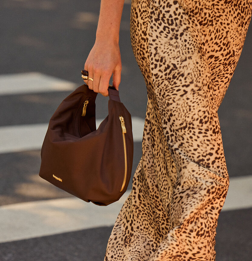 Close-up of a woman carrying a chocolate brown Baggallini hobo bag with gold hardware while wearing a leopard print skirt.