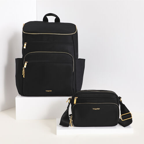 Baggallini black twill handbags featuring a backpack and crossbody bag with gold-tone hardware displayed on white pedestals from the Manhattan collection.