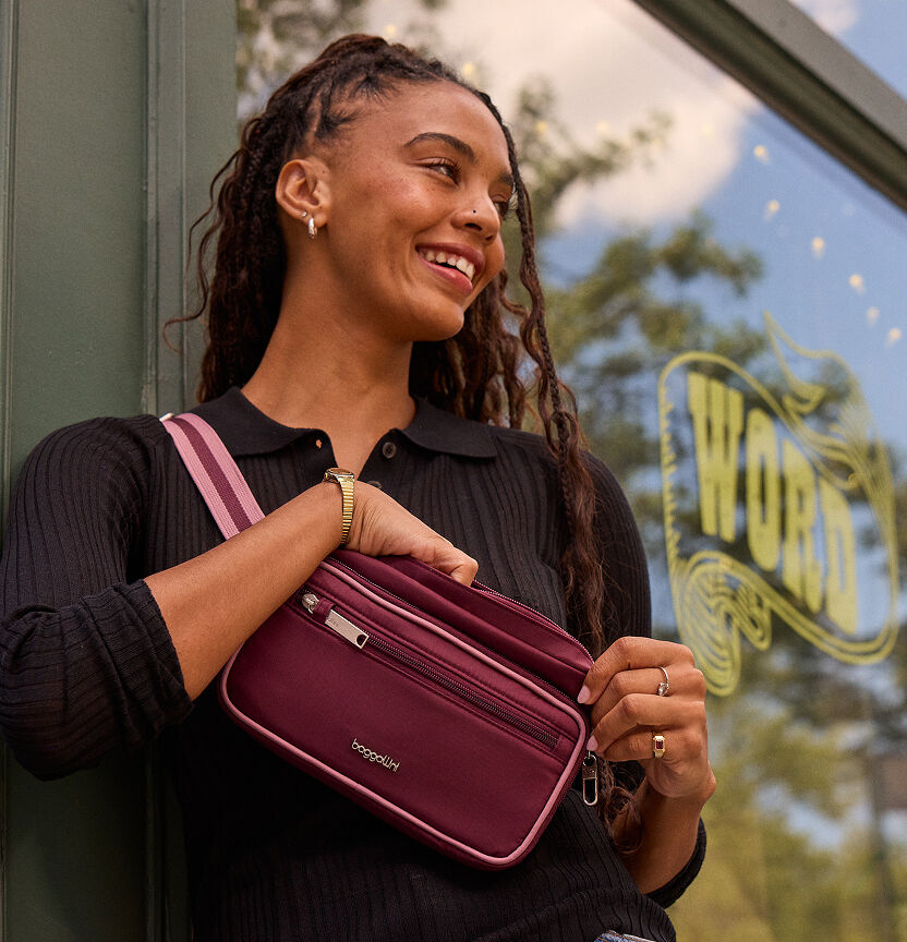 Woman wearing a burgundy Baggallini crossbody bag with multiple zip pockets while standing outside a coffee shop.
