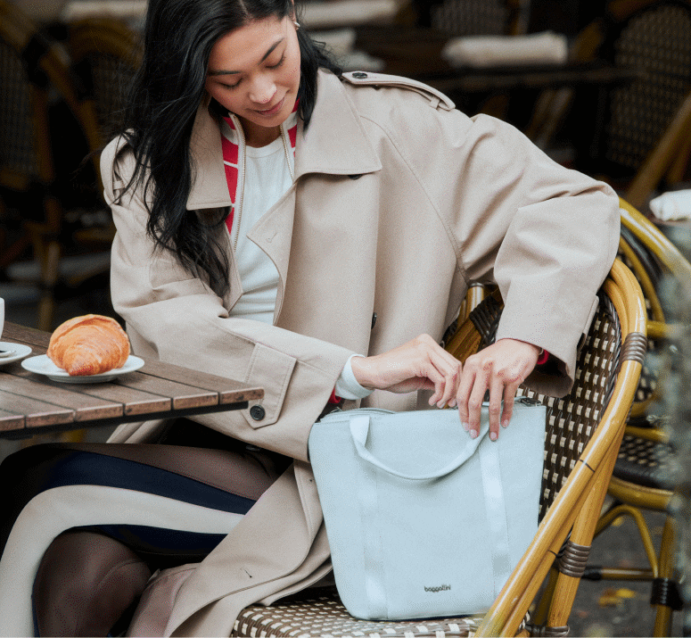 Woman in a beige coat sits at an outdoor caf&eacute; table with a croissant, placing something into a light gray tote bag on her lap.