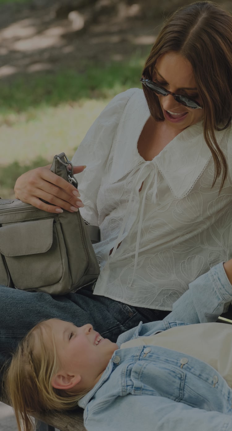Woman sitting on a park bench with baggallini bag on her lap, smiling at a young child lying across the bench with legs resting on her lap. Trees and grass are visible in the background.