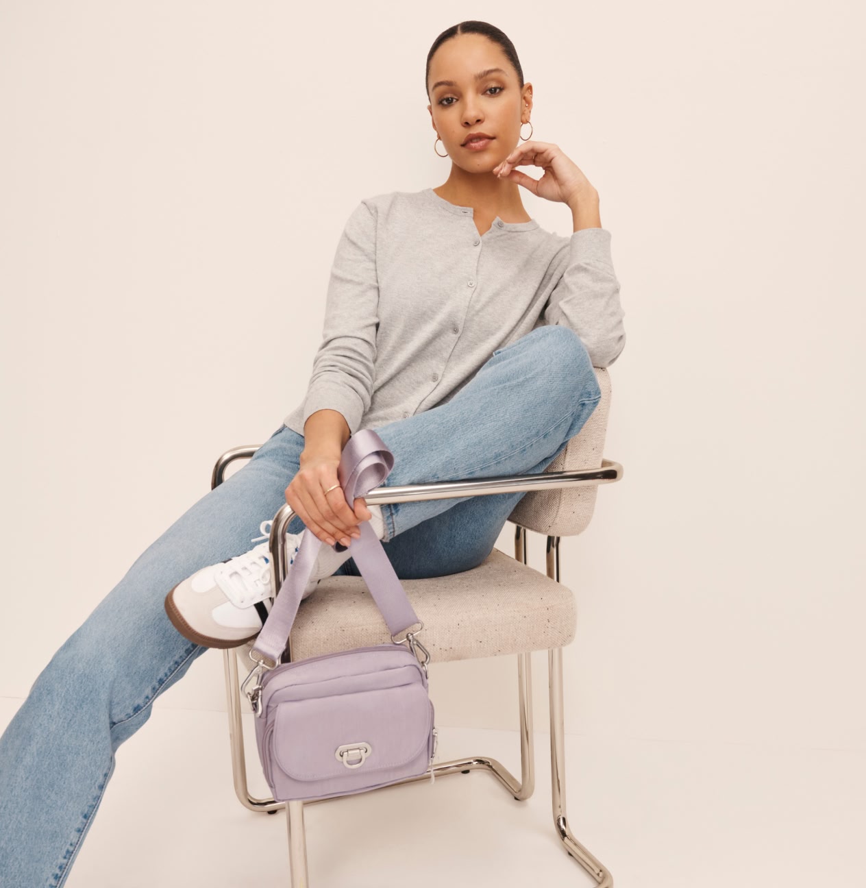 Woman seated in a modern chair wearing a gray cardigan, light-wash jeans, and sneakers while holding a lavender crossbody bag with a wide strap.