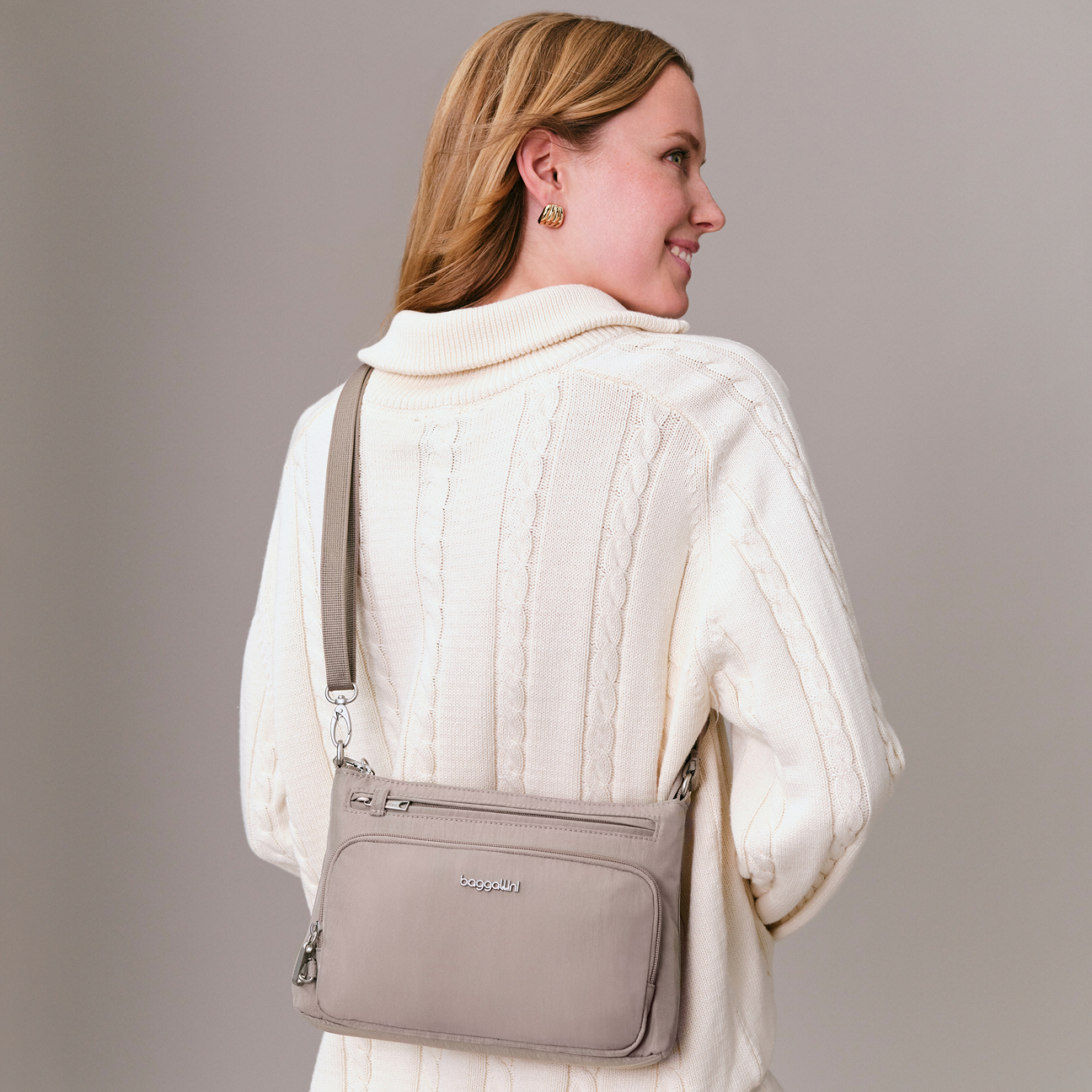 Woman wearing a cream cable-knit sweater and gold earrings, carrying a light beige crossbody bag with zippered pockets.
