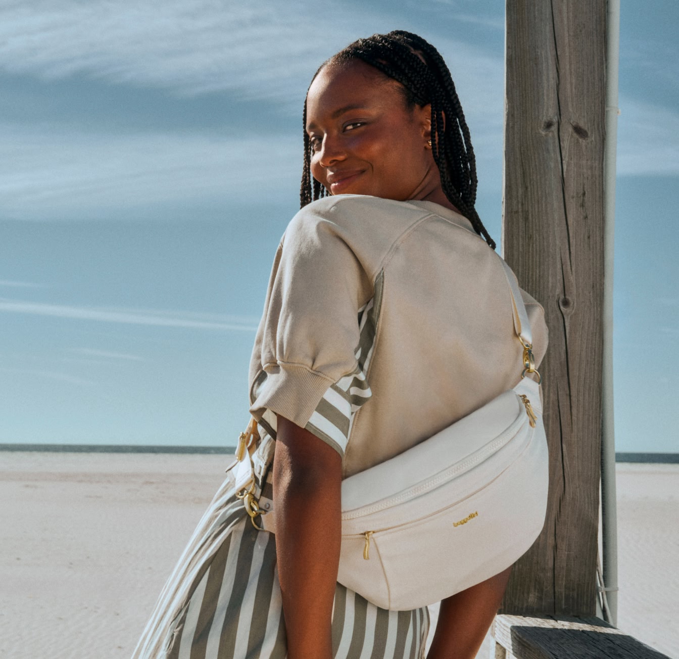 A woman stands on a sandy beach, smiling over her shoulder while wearing a beige top, striped skirt, and a white Manhattan crossbody bag.