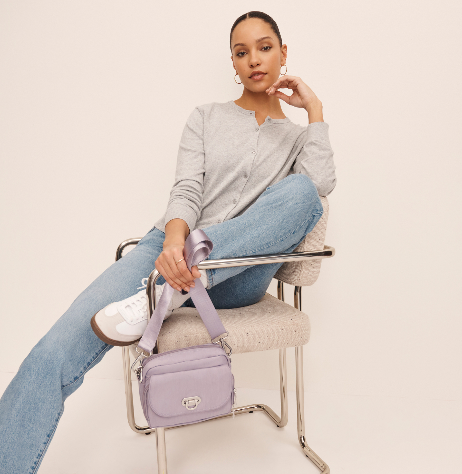 Woman seated in a modern chair wearing a gray cardigan, light-wash jeans, and sneakers while holding a lavender crossbody bag with a wide strap.