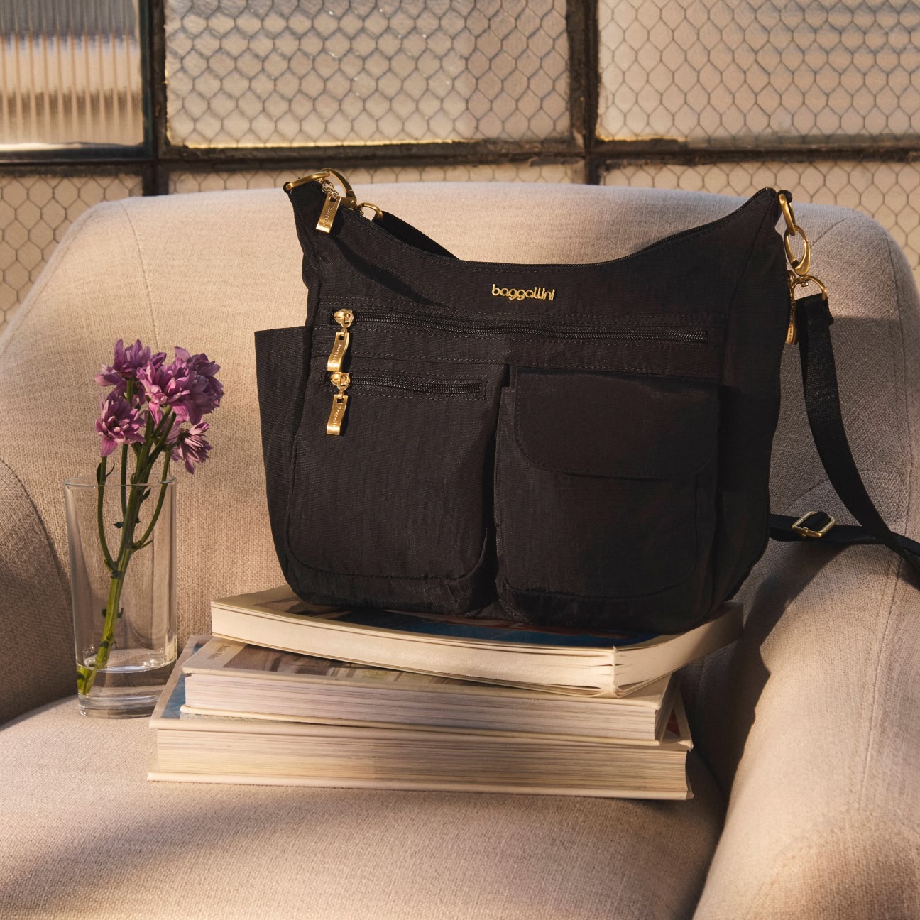 A black baggallini shoulder bag with gold zippers sits on a stack of books on a beige chair next to a glass of purple flowers.